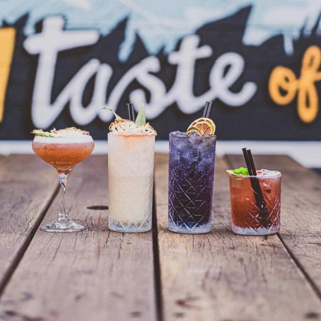 Four colorful cocktails lined up on a wooden table, each in a different glass and garnished with fruit, including citrus slices and pineapple. The drinks range in color from light pink and creamy white to deep purple and amber.
