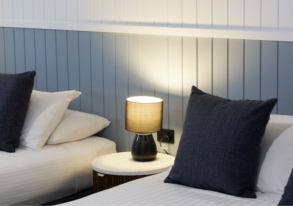 Close-up of twin beds in a Deluxe Queen Room at Settlers Inn Port Macquarie, with white bedding, dark cushions and bedside lamp.