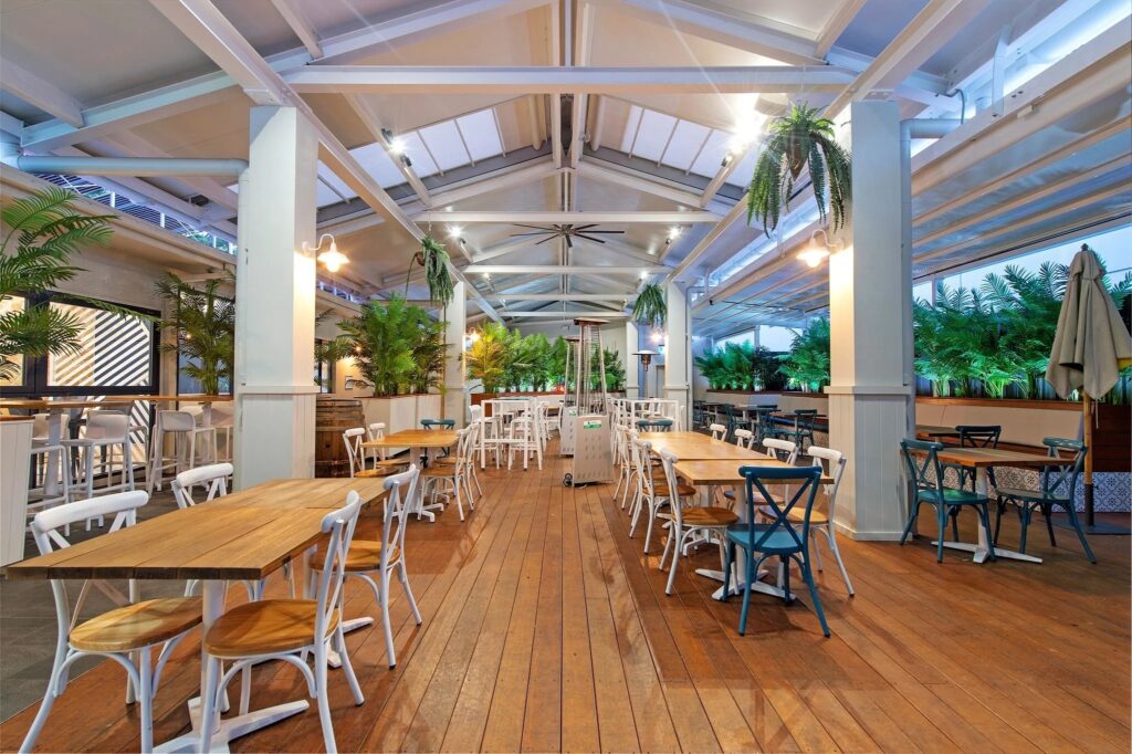A bright, open pavilion-style dining space with a high white ceiling and exposed beams. Wooden floors are lined with neatly arranged tables and white chairs, while lush green plants border the area. Soft lighting and heaters create a warm, inviting atmosphere suitable for functions or group dining.