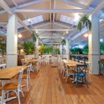 A bright, open pavilion-style dining space with a high white ceiling and exposed beams. Wooden floors are lined with neatly arranged tables and white chairs, while lush green plants border the area. Soft lighting and heaters create a warm, inviting atmosphere suitable for functions or group dining.